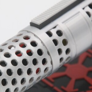 May include: A cylindrical object with a white perforated outer shell featuring a repeating circular hole pattern. A rectangular gray switch is mounted on the side. Through the perforations, a red internal component is visible. A red emblem is nearby.