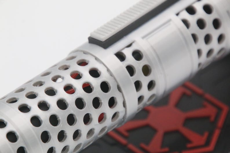 May include: A cylindrical object with a white perforated outer shell featuring a repeating circular hole pattern. A rectangular gray switch is mounted on the side. Through the perforations, a red internal component is visible. A red emblem is nearby.