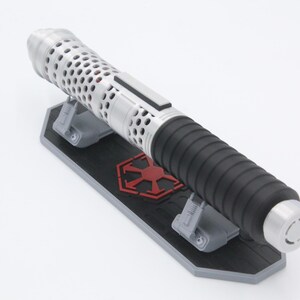 May include: A silver and black lightsaber hilt featuring a perforated upper section and a ribbed grip, resting horizontally on a black and gray display stand that displays a red imperial-style emblem on its central surface.