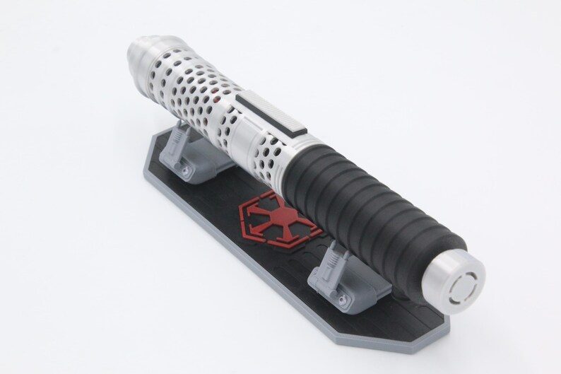 May include: A silver and black lightsaber hilt featuring a perforated upper section and a ribbed grip, resting horizontally on a black and gray display stand that displays a red imperial-style emblem on its central surface.