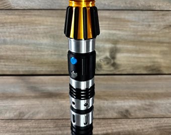 May include: A silver, black, and gold lightsaber hilt. The hilt features a ribbed silver top and bottom, with a black and gold midsection. A blue button is visible on the black section. The lightsaber is cylindrical and has a detailed design.