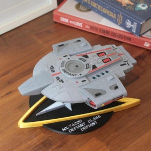 May include: A grey and red Star Trek starship model of the USS Defiant, NX-74205, on a black base with a yellow Starfleet insignia. The model is labelled "USS Defiant" and "NX-74205 Defiant Class Defiant".