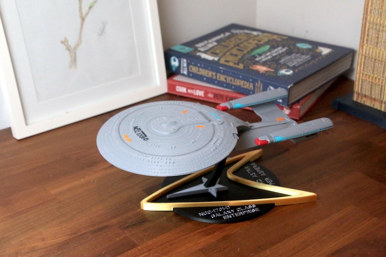 K&ouml;nnte beinhalten: Ein graues Modell des Raumschiffs USS Enterprise NCC-1701-D aus Star Trek: The Next Generation, auf einem schwarzen St&auml;nder mit goldener Verzierung. Der St&auml;nder tr&auml;gt den Text "NCC-1701-D GALAXY CLASS ENTERPRISE".