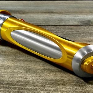 May include: A gold and silver lightsaber prop. The hilt is primarily gold with silver accents and a central silver panel. The design features a cylindrical shape with detailed end caps and a button.