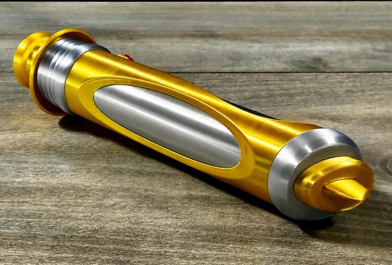 May include: A gold and silver lightsaber prop. The hilt is primarily gold with silver accents and a central silver panel. The design features a cylindrical shape with detailed end caps and a button.