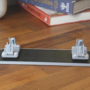 May include: A gray and black 3D-printed object, possibly a model or display stand. It features a rectangular base with a textured black surface and two gray structures at each end. The object is set on a wooden surface.