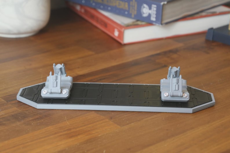 May include: A gray and black 3D-printed object, possibly a model or display stand. It features a rectangular base with a textured black surface and two gray structures at each end. The object is set on a wooden surface.