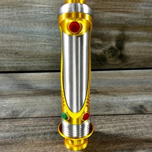 May include: A metallic silver and gold lightsaber prop with red and green button accents. The hilt features a gold base and top, with a silver center section. The design includes a series of gold accents and a red button.