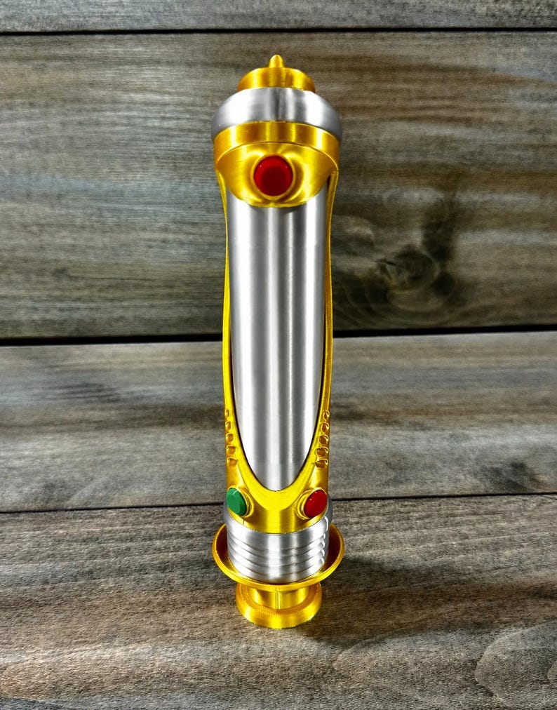 May include: A metallic silver and gold lightsaber prop with red and green button accents. The hilt features a gold base and top, with a silver center section. The design includes a series of gold accents and a red button.