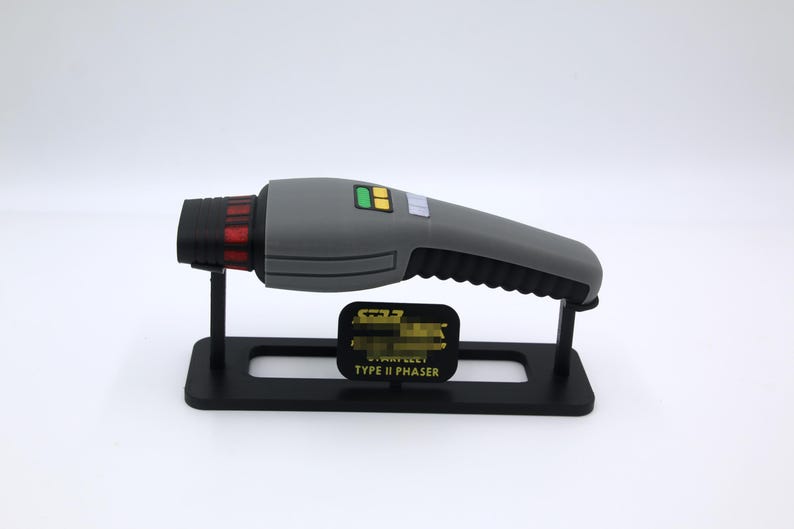 K&ouml;nnte beinhalten: Eine graue und schwarze Starfleet Type II Phaser-Replik auf einem schwarzen Display-St&auml;nder. Der Phaser hat einen roten und schwarzen Lauf und ein gelbes und gr&uuml;nes Bedienfeld.