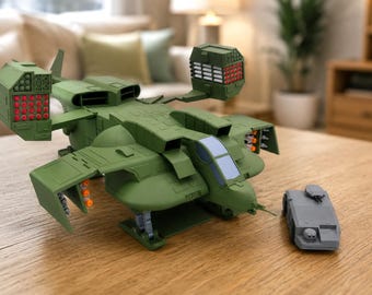 May include: A matte olive green plastic dropship toy featuring two large elevated missile pods with red tips, a central cockpit with a light blue window, and side-mounted orange projectiles. A small gray armored vehicle sits beside the aircraft.