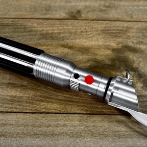 May include: A silver and black lightsaber prop with a red button and a curved blade. The lightsaber is lying on a wooden surface.