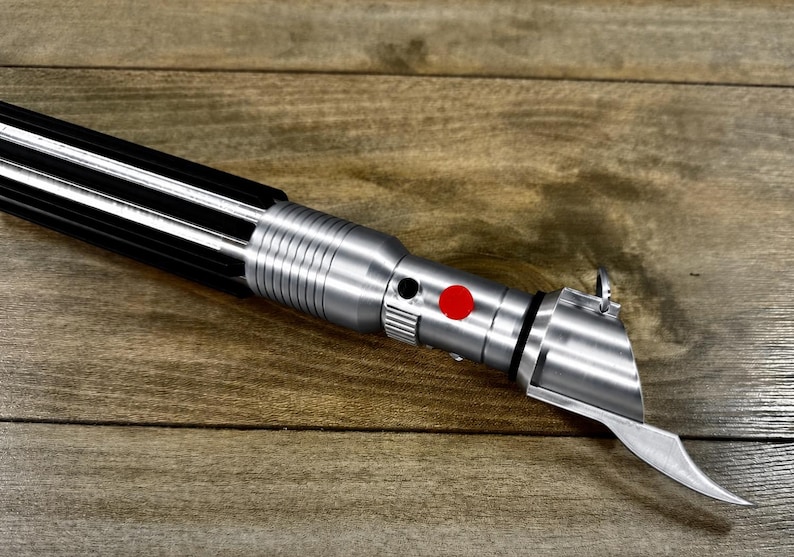 May include: A silver and black lightsaber prop with a red button and a curved blade. The lightsaber is lying on a wooden surface.