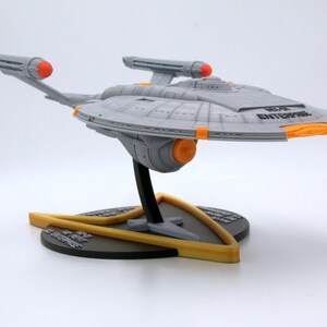 Puede incluir: Modelo gris de la nave espacial U.S.S. Enterprise, con detalles en naranja y amarillo, montado sobre un soporte negro y amarillo. La nave tiene el texto "NCC-1701 ENTERPRISE".