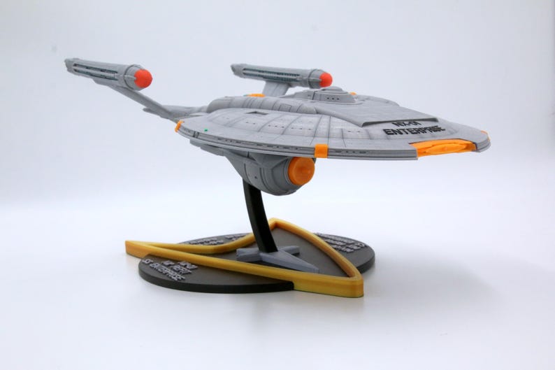 Puede incluir: Modelo gris de la nave espacial U.S.S. Enterprise, con detalles en naranja y amarillo, montado sobre un soporte negro y amarillo. La nave tiene el texto "NCC-1701 ENTERPRISE".