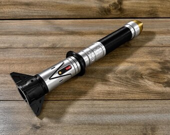 May include: A silver and black lightsaber prop with a ribbed black handle and a gold-colored tip. The hilt features a red and yellow button and a black triangular emitter. The prop is lying on a wooden surface.