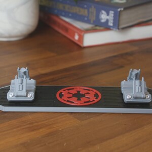 May include: A black and gray Star Wars-themed desk organizer with two gray vehicle models and a red Imperial symbol. The organizer is rectangular with angled sides and sits on a wooden surface.