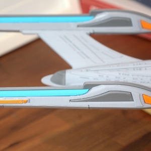 Puede incluir: Un modelo blanco y gris de una nave espacial con detalles azules y naranjas. La nave espacial tiene un cuerpo largo y estrecho con una nariz puntiaguda y dos alas. Las alas tienen una franja azul que recorre la parte superior y una peque&ntilde;a franja naranja que recorre la parte inferior.