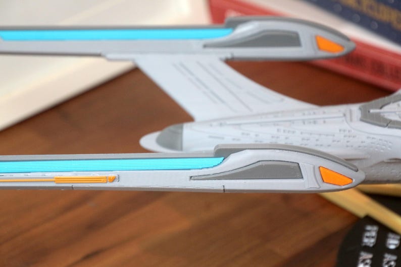 Puede incluir: Un modelo blanco y gris de una nave espacial con detalles azules y naranjas. La nave espacial tiene un cuerpo largo y estrecho con una nariz puntiaguda y dos alas. Las alas tienen una franja azul que recorre la parte superior y una peque&ntilde;a franja naranja que recorre la parte inferior.