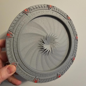 May include: A gray, circular, 3D-printed model of a Stargate. The outer ring features detailed carvings and red accents. The center has a spiral design with a star-shaped element. The model is held by a hand.