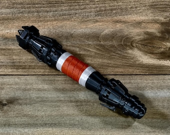 May include: A black and silver lightsaber prop with a red wrapped grip. The prop is lying on a wooden surface. The lightsaber has intricate details and a futuristic design, perfect for cosplay or display.