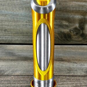 May include: A gold and silver decorative bottle stopper. The stopper has a cylindrical body with an oval silver insert. The top and base are gold, with a silver band in between. The top has a small, gold, stylized design.