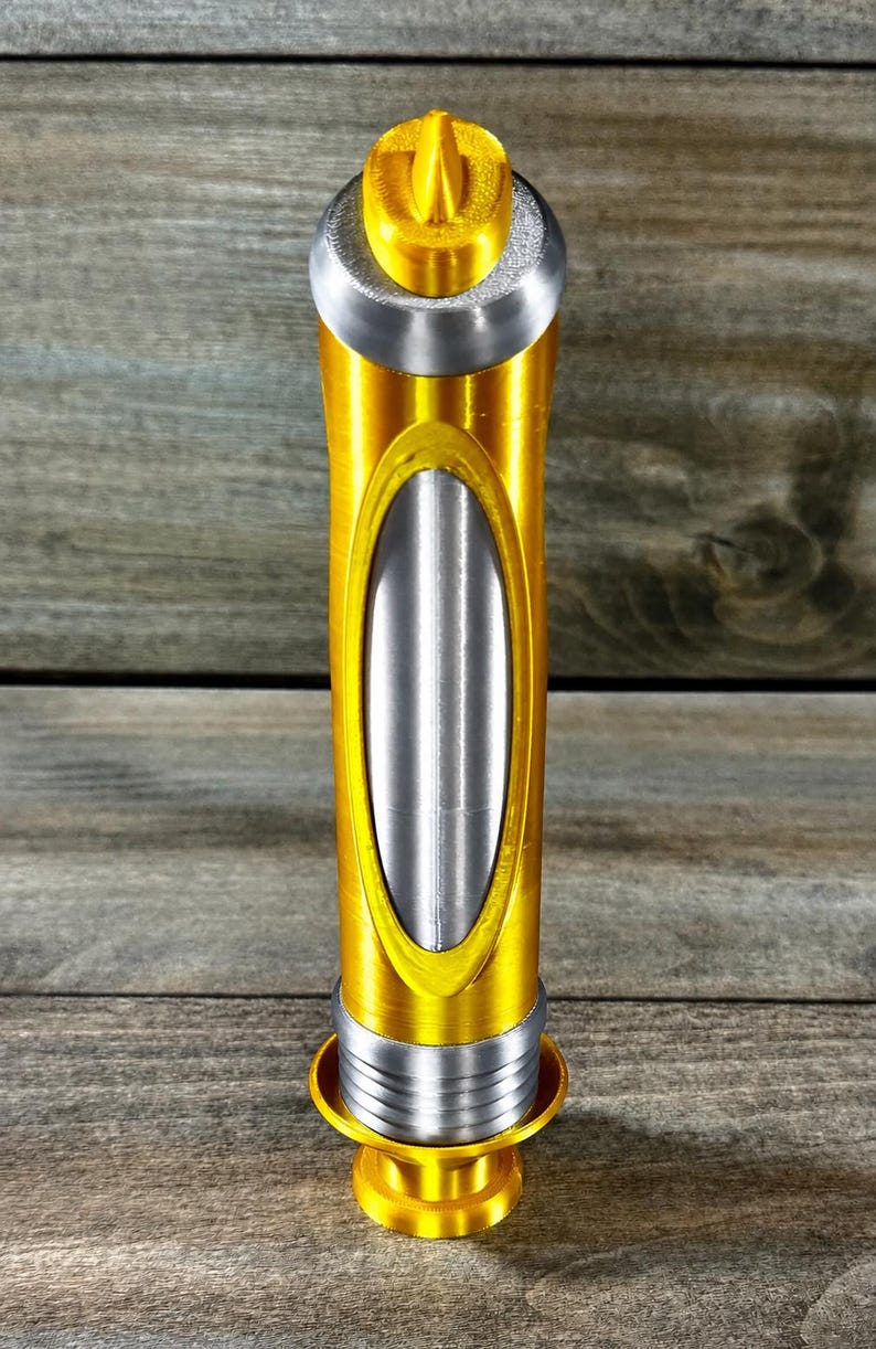 May include: A gold and silver decorative bottle stopper. The stopper has a cylindrical body with an oval silver insert. The top and base are gold, with a silver band in between. The top has a small, gold, stylized design.