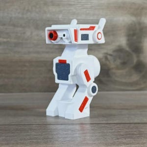 May include: A white robot toy with red and gray accents. The toy has a cylindrical head with a red eye and a rectangular body. Red stripes and gray squares are placed on the body. The toy is standing on a wooden surface.
