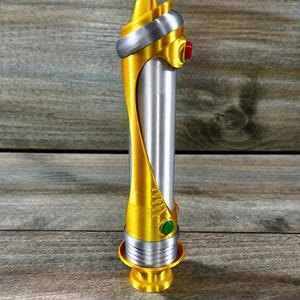 May include: A gold and silver lightsaber prop with a cylindrical silver body and gold accents. The hilt features a red and green button. The lightsaber rests on a gold base.