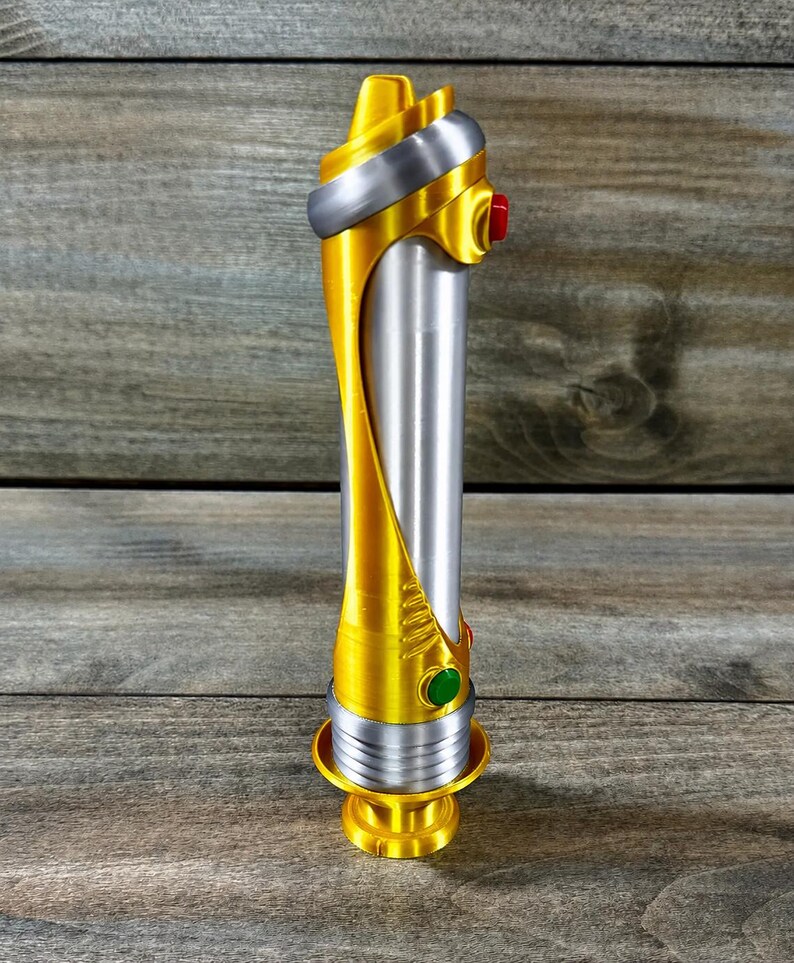 May include: A gold and silver lightsaber prop with a cylindrical silver body and gold accents. The hilt features a red and green button. The lightsaber rests on a gold base.