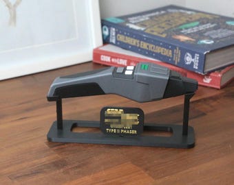 May include: A gray Star Trek Type II Phaser replica on a black display stand with a gold plaque that reads "STAR TREK TYPE II PHASER".