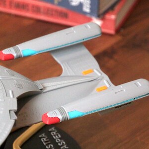 K&ouml;nnte beinhalten: Ein graues und wei&szlig;es Modell eines Star Trek Raumschiffs mit roten und blauen Akzenten. Das Schiff hat einen Untertassenbereich und zwei Triebwerke. Das Schiff steht auf einem schwarzen Sockel mit der Aufschrift "Astra Aspera".