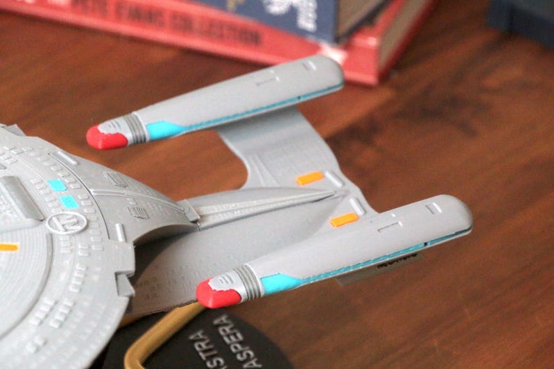 K&ouml;nnte beinhalten: Ein graues und wei&szlig;es Modell eines Star Trek Raumschiffs mit roten und blauen Akzenten. Das Schiff hat einen Untertassenbereich und zwei Triebwerke. Das Schiff steht auf einem schwarzen Sockel mit der Aufschrift "Astra Aspera".