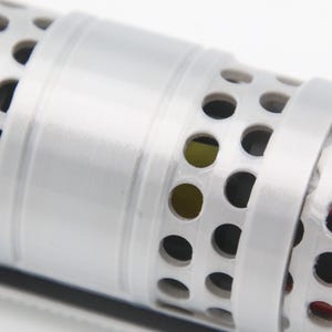 May include: A cylindrical object featuring a white exterior with a repeating pattern of circular cutouts. The surface is divided by a smooth, solid white band that encircles the center. Through the perforations, glimpses of red and yellow inner components are visible.