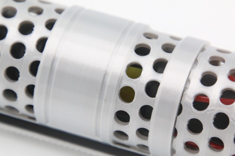 May include: A cylindrical object featuring a white exterior with a repeating pattern of circular cutouts. The surface is divided by a smooth, solid white band that encircles the center. Through the perforations, glimpses of red and yellow inner components are visible.