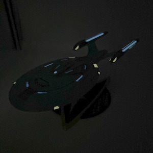 Puede incluir: Un modelo de nave espacial gris oscuro con una base negra. La nave espacial tiene una forma de platillo redondeado con una nariz puntiaguda y dos alas. El modelo est&aacute; iluminado con luces azules y amarillas.