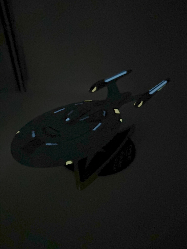 Puede incluir: Un modelo de nave espacial gris oscuro con una base negra. La nave espacial tiene una forma de platillo redondeado con una nariz puntiaguda y dos alas. El modelo est&aacute; iluminado con luces azules y amarillas.