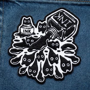 May include: Black and white embroidered patch featuring a whimsical design of ink spilling from a bottle, forming cat-like shapes. The word "INK" is visible on the ink bottle. The patch is set against a denim background.