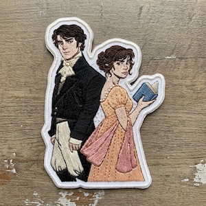 May include: Embroidered patch featuring a man and woman in period clothing. The man wears a black coat and white cravat, while the woman holds a blue book and wears a peach-colored dress with pink accents. The patch has a white border.