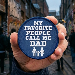 May include: A round, blue patch with white embroidered text that reads "MY FAVORITE PEOPLE CALL ME DAD." Below the text is a white silhouette of a family. The patch is held in a hand, with a blurred urban background.