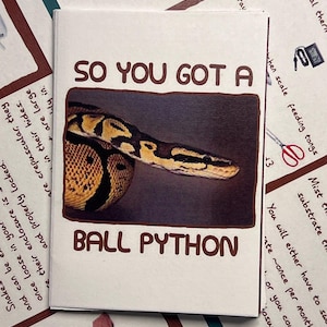 May include: A white card with the text "SO YOU GOT A BALL PYTHON" in brown lettering. The card features a photo of a ball python snake with a yellow and brown pattern.