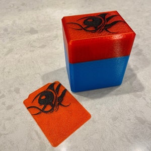 May include: A red and blue 3D printed box with a black design on the top. The box is open and the lid is laying on the table. The lid is also red and has the same black design.