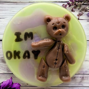 May include: A round, light green soap with a brown teddy bear design and the words "I'M OKAY" in dark brown. The bear has a stitched appearance and is set against a marbled, pastel background.