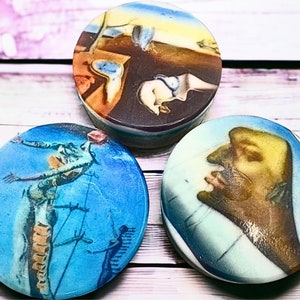 May include: Three round ceramic coasters featuring famous Salvador Dali paintings. The first coaster features a melting clock, the second features a figure in a blue sky, and the third features a close-up of a face.