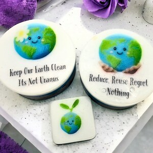 May include: Three round white soap bars with a blue and green earth design and a smiling face. The soap bars have the following text: "Keep Our Earth Clean Its Not Uranus", "Reduce, Reuse Regret Nothing", and a small green sprout.