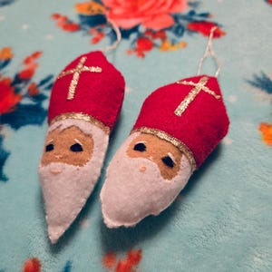 May include: Two felt ornaments shaped like Saint Nicholas, each with a red hat, white beard, and a gold cross.