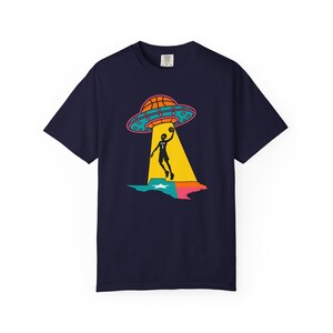 May include: Navy blue t-shirt featuring a basketball-themed graphic. The design includes a basketball, a UFO, and a basketball player silhouetted in a beam of light over a colorful map. The shirt is a short-sleeved crew neck.