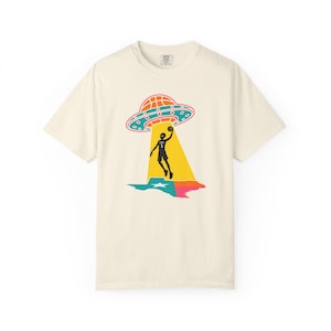 May include: Cream-colored t-shirt featuring a graphic of a basketball player being beamed up by a UFO. The UFO is orange and teal, and the player is silhouetted in black. The design includes a colorful Texas outline.