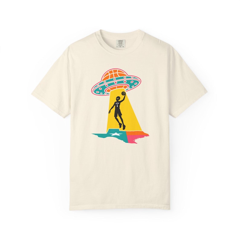 May include: Cream-colored t-shirt featuring a graphic of a basketball player being beamed up by a UFO. The UFO is orange and teal, and the player is silhouetted in black. The design includes a colorful Texas outline.
