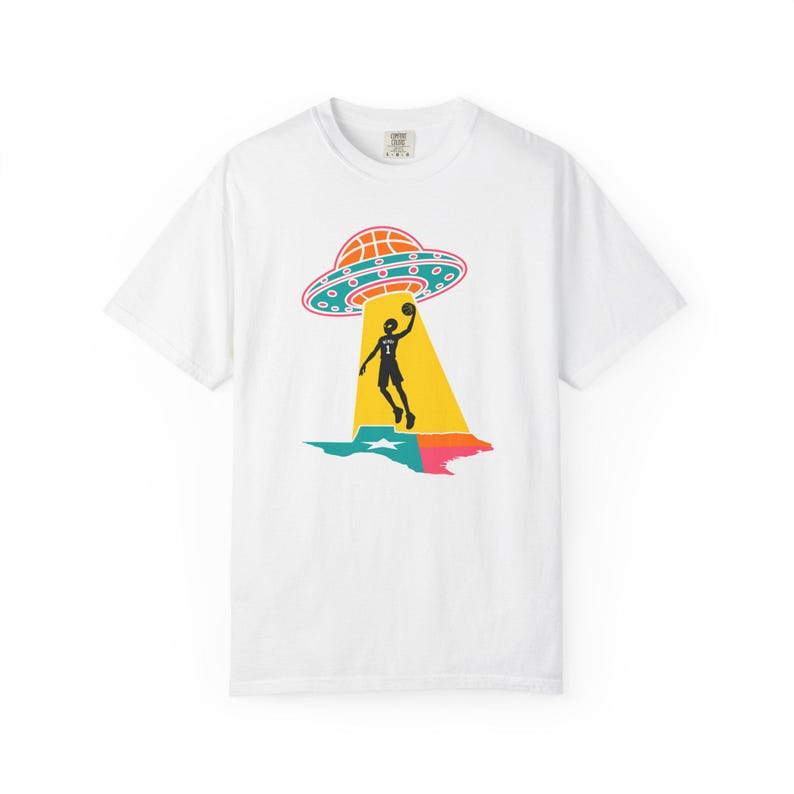 May include: White t-shirt featuring a graphic of a basketball player being beamed up by a UFO, with the state of Texas in the background. The UFO is orange and teal, and the player is black. The Texas outline is teal, pink, and yellow.